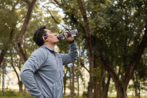 Thirsty runner man is drinking water bottle with earphone after exercise cardio on path in forest background.