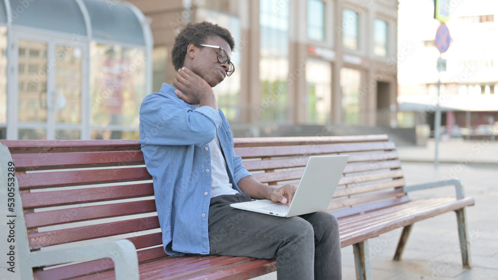 Young African Man with Neck Pain Using Laptop while Sitting Outdoor on Bench Young African Man with Neck Pain Using Laptop while Sitting Outdoor on Bench
