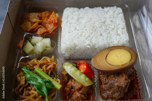 Serving rice boxes in white boxes from cardboard or paper boxes. White rice, potato fried chili sauce, pickled cucumber, vegetables, egg bacem, beefsteak beef rendang. Potato chips, chili paste.  photo