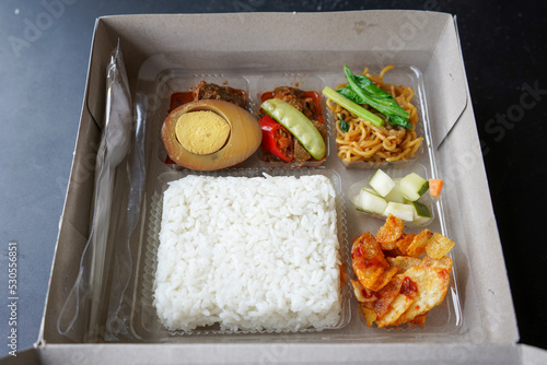 Serving rice boxes in white boxes from cardboard or paper boxes. White rice, potato fried chili sauce, pickled cucumber, vegetables, egg bacem, beefsteak beef rendang. Potato chips, chili paste.  photo