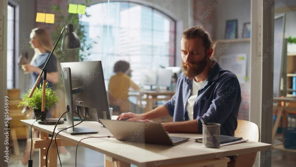 Stylish Long-Haired Bearded Specialist Sitting at a Desk in Creative Agency. Young Stylish Man Working on a Laptop Computer Connected to a Second Display. Office Team Members in the Background. Stylish Long-Haired Bearded Specialist Sitting at a Desk in Creative Agency. Young Stylish Man Working on a Laptop Computer Connected to a Second Display. Office Team Members in the Background.
