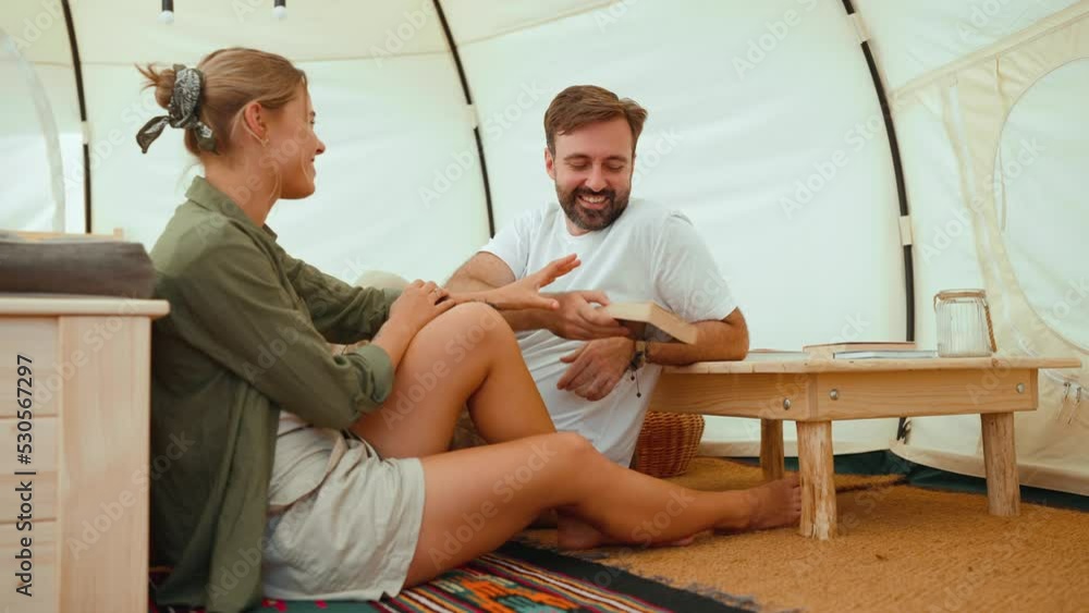 Laughing blond young couple reading book and talking while sitting in a tent Laughing blond young couple reading book and talking while sitting in a tent