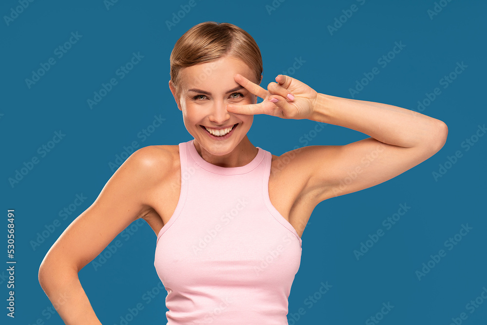 Optimistic energized smiling blond woman with short haircut showing peace sign, posing on blue studio background. Joyful mood. Optimistic energized smiling blond woman with short haircut showing peace sign, posing on blue studio background. Joyful mood.