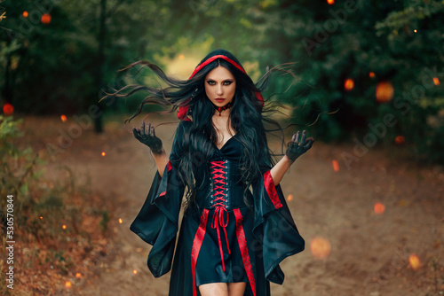 Fantasy woman witch creates magic, hands raised long hair flying in wind motion. Sexy face dark eyes look at camera. summer nature green forest trees. Gothic girl art black red dress old vintage style