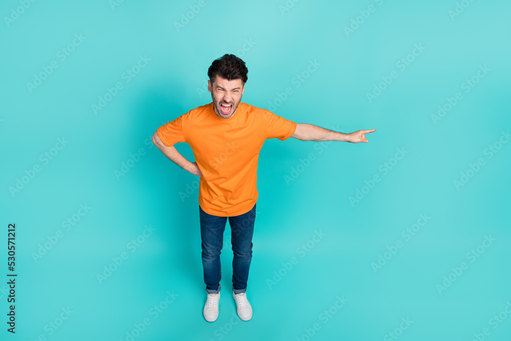 Full length photo of young funny handsome agressive man wear orange t-shirt denim jeans finger pointing empty space angry chief isolated on cyan color background Full length photo of young funny handsome agressive man wear orange t-shirt denim jeans finger pointing empty space angry chief isolated on cyan color background