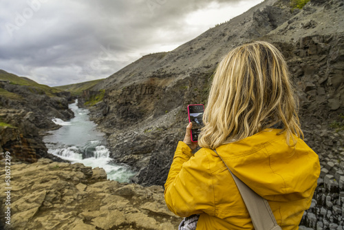 Donna bionda in viaggio mentre fotografa con un cellulare un canyon con un fiume in Islanda photo