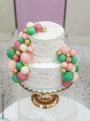 Vem pra festa e comemore. Um bolo de aniversário de dois andares, enfeitado com bolas verdes e rosa  sob um pedestal dourado e um suporte de acrilico. photo