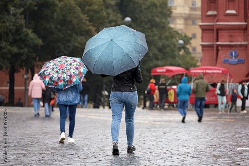 Rain in city, people with umbrellas walk on a street, woman in jeans in foreground. Rainy weather in autumn