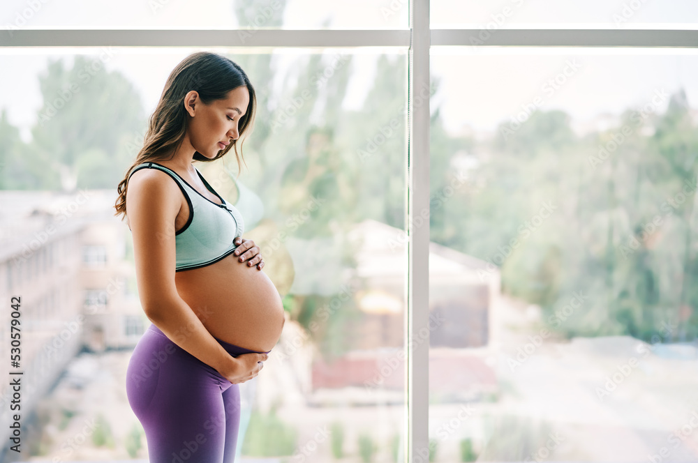 Portrait of young pregnant woman in sportswear standing near window holding hands on her belly with copy space