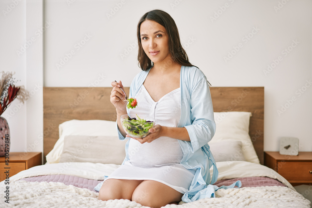 Portrait of young pregnant woman in pajama eating a fresh salad sitting in bed Portrait of young pregnant woman in pajama eating a fresh salad sitting in bed