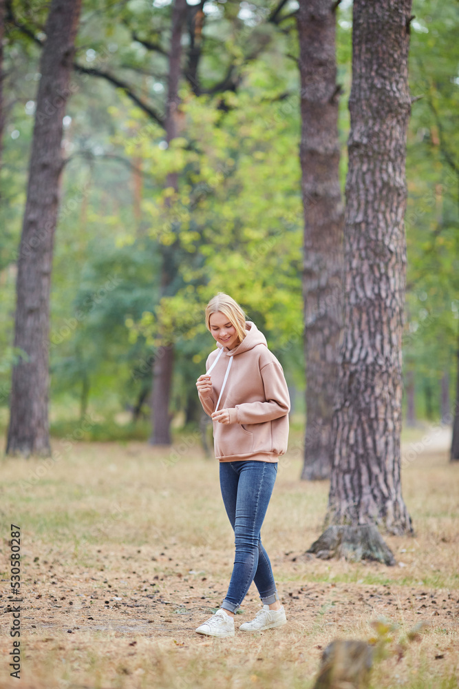 Blue-eyed blonde in a beige hoodie walks in a pine forest. Portrait of a joyful young woman enjoying in autumn park. Blue-eyed blonde in a beige hoodie walks in a pine forest. Portrait of a joyful young woman enjoying in autumn park.