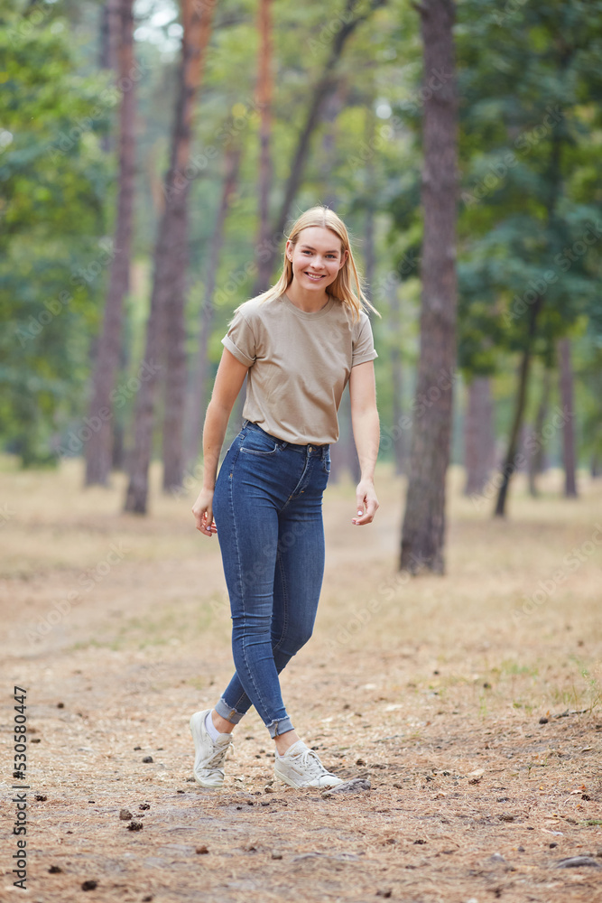 Blue-eyed blonde in a beige T-shirt walks in a pine forest. Portrait of a joyful young woman enjoying in autumn park.