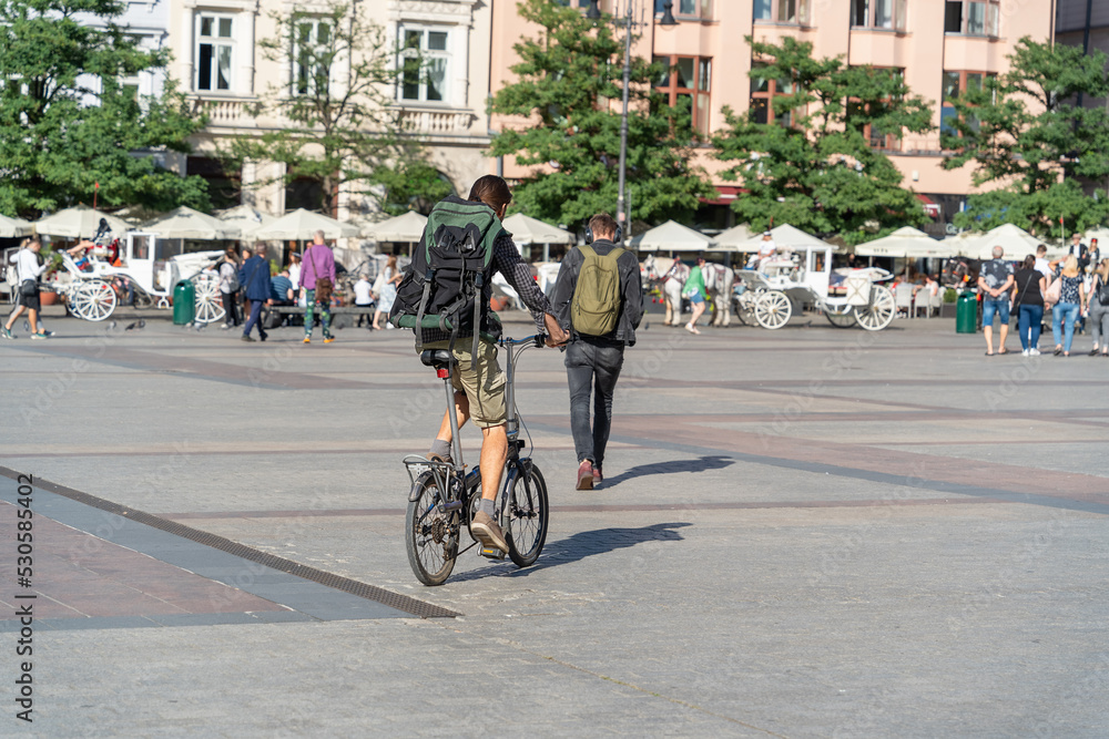 A tourist with a travel backpack on his back rides a bike on a sunny day in the city square behind a backpacker with a white horse-drawn carriage in the background, the market square in Krakow