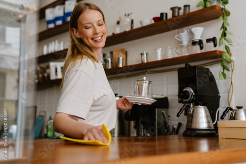 Blonde white barista woman smiling while cleaning counter in cafe