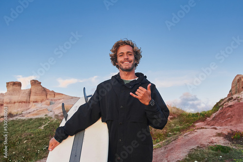 Cheerful positive man surfer carrying his surfboard and showing shaka gesture to the camera photo