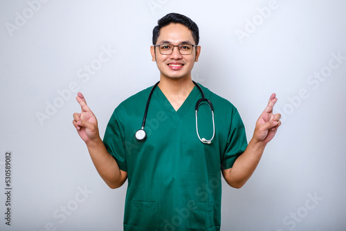 Handsome asian male wearing green scrubs uniform gesturing finger crossed smiling with hope and eyes closed. luck and superstitious concept