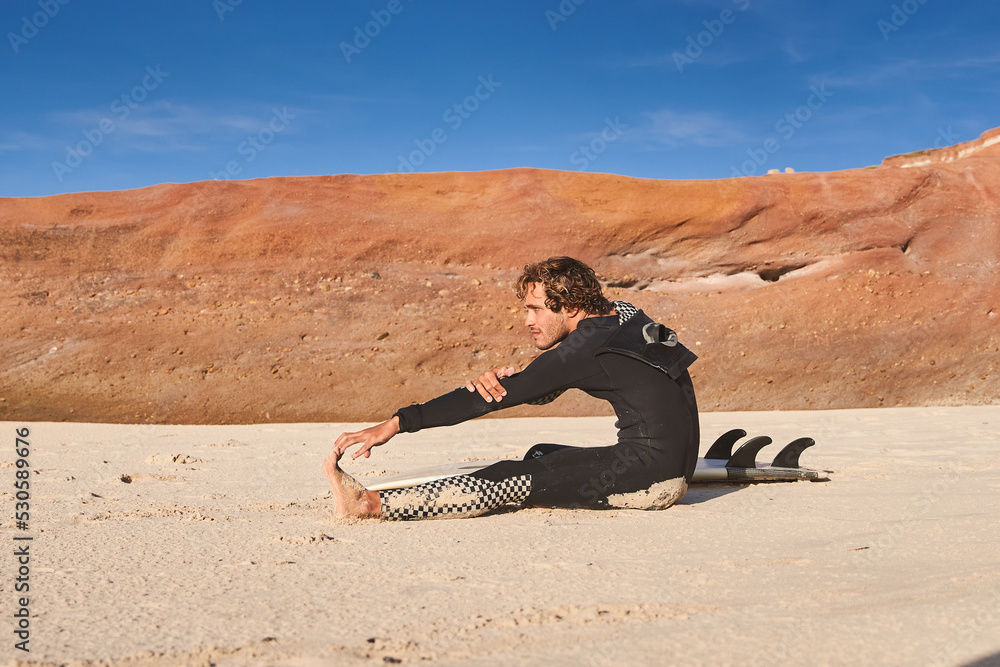 Caucasian man sitting at the sand in front of the sea and stretching