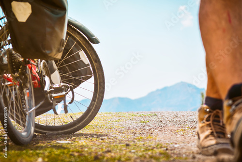 Close up tracking low angle back view of male cyclist walking towards away touring bicycle isolated in scenic road panorama background. bicycle touring and getting fit concept