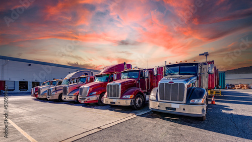 Lined up Semi trucks on a parking lot at logistics warehouse with orange sunset sky
