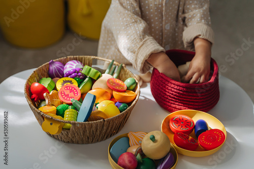 Fototapeta Naklejka Na Ścianę i Meble -  A little girl playing with  wooden fruits and vegetables on the table in nursery. Educational games, learning through play. Developing Montessori toddlers activities.