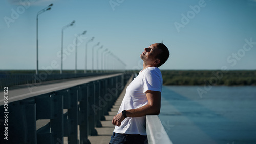 A man in a white T-shirt and jeans stands on a bridge across the river and enjoys life, raising his face to the sun. A man demonstrates mental balance and health