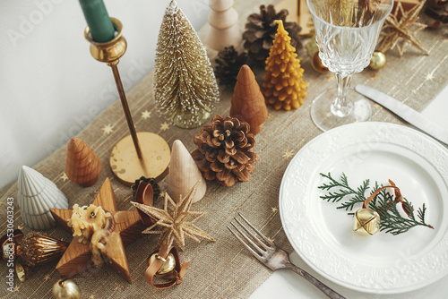 Stylish Christmas table setting. Fir branch with bell on plate, vintage cutlery, wineglass, modern golden christmas trees and candles on  rustic table. Holiday brunch, new year celebration