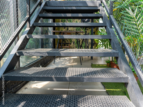 Black steel staircase with regular tread plate pattern for slip resistance, metal webbing, and greenery decoration on the right.
