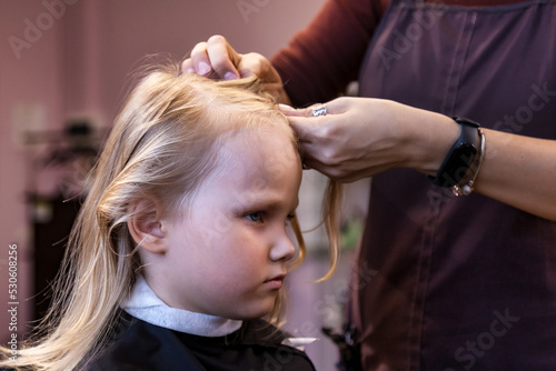 Barber woman make fashionable braids hairstyle for cute little blond girl child in modern barbershop, hair salon. Hairdresser makes hairdo for young baby in barber shop. Concept hairstyle, beauty