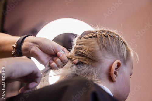 Barber woman make fashionable braids hairstyle for cute little blond girl child in modern barbershop. Hair salon, Hairdresser makes hairdo for baby in barber shop close up. Concept hairstyle, beauty