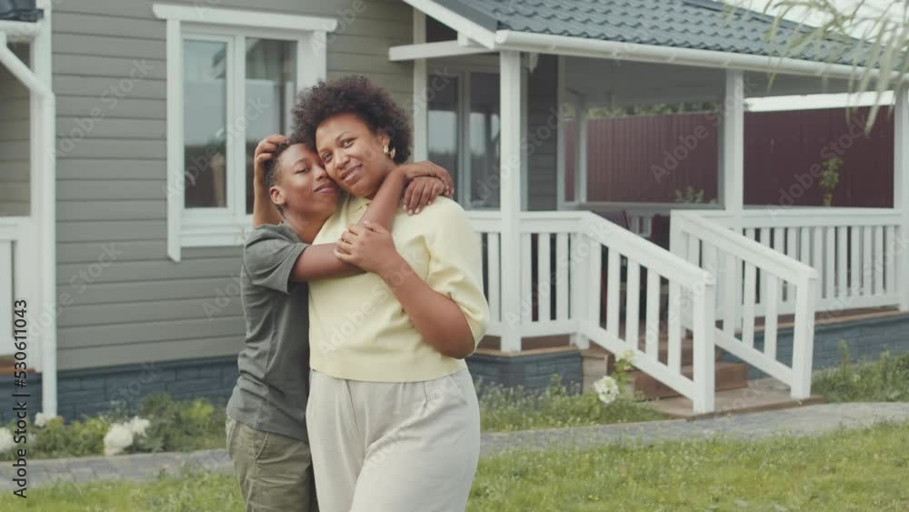 Medium portrait of delighted African American woman and her loving 12 year old son posing for camera standing outside their house in summer Medium portrait of delighted African American woman and her loving 12 year old son posing for camera standing outside their house in summer