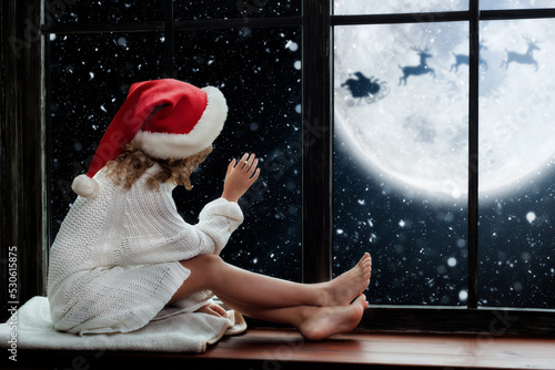 Merry Christmas. Cute little child sitting by window and looking at Santa Claus flying in his sleigh against moon sky. Happy kid enjoying holiday.