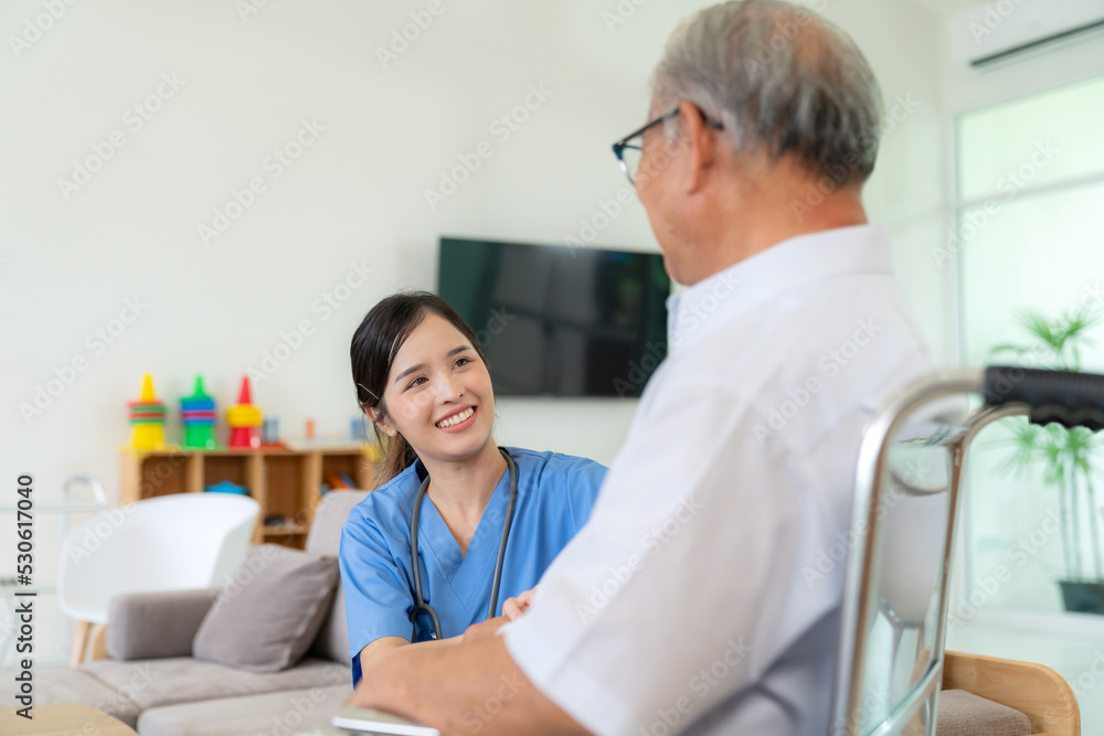 Obraz premium Young Asia caregiver doctor sitting beside elderly man who isit on wheelchair in the livingroom at nursing home. Mental care and wellness. Nurse taking care old man and physical therapy with smile