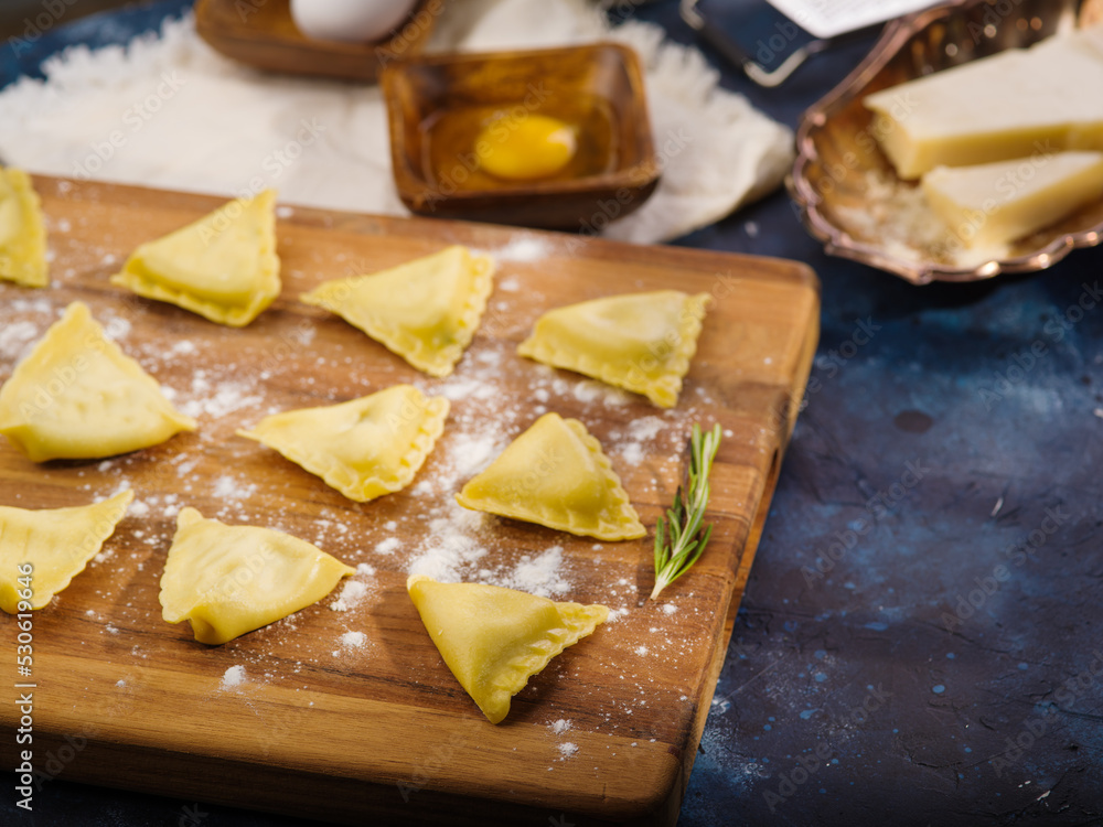 Triangular raw Italian ravioli on a floured wooden cutting board. In the background are ingredients and kitchen utensils on a dark blue background. Recipes for restaurant and home cooking.
