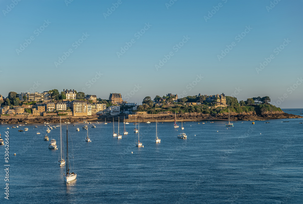 St. Malo, Brittany, France - July 8, 2022: Morning light over Rance river mouth to Pointe du Moulinet on peninsula with its many buildings under blue sky. English Channel behind. Yachts up front