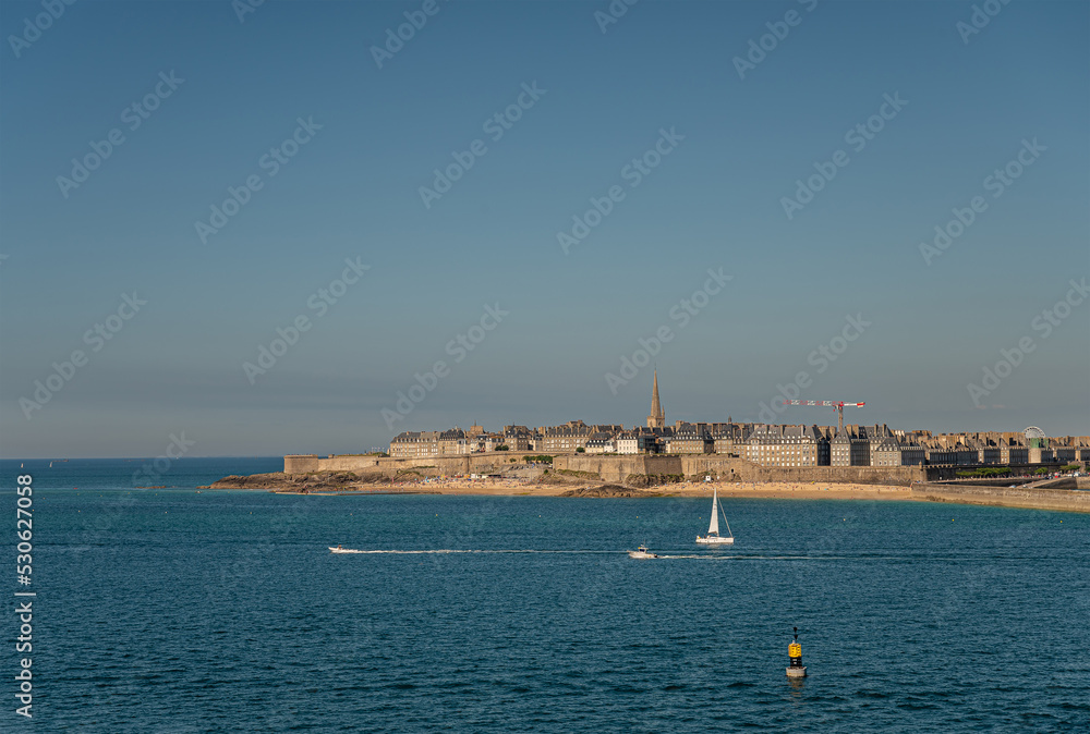 St. Malo, Brittany, France - July 8, 2022: Facing English Channel, Historic old town with its tall palaces behind its ramparts, under blue sky. Blue water up front. Spire of Cathedral, Yachts on water