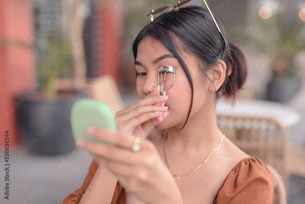 A beautiful Asian woman uses a pressed powder mirror to help curl her long eyelashes with an eyelash curler.