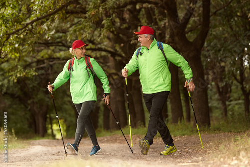 Married couple, happy middle age woman and man walking with Scandinavian sticks in autumn forest, outdoors. Nordic walking, healthy lifestyle