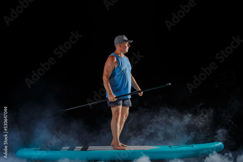 A man in a cap on a sub board with a paddle on a black background in the fog.