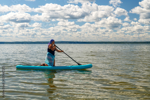 a woman in a closed swimsuit, a pareo and a headdress on her knees on a SUP board with an oar floats on the water.