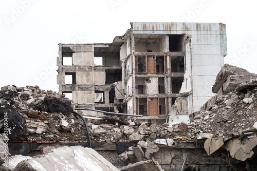 A ruined building against a gray sky with piles of concrete rubble and construction debris in the foreground. Background