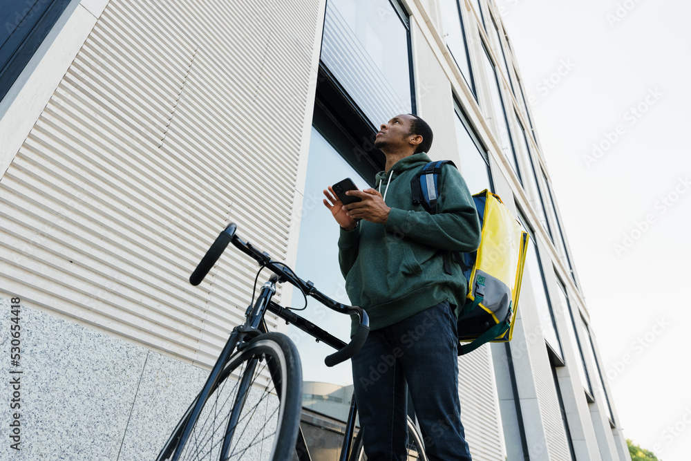 Portrait of a delivery guy with a backpack standing next to a bicycle and looking at an office building. The courier is holding a smartphone. Lunch delivery.