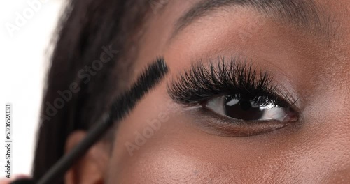 Close up view of a black woman eye. Applying mascara using a brush to her eyelashes. Isolated on a white background.