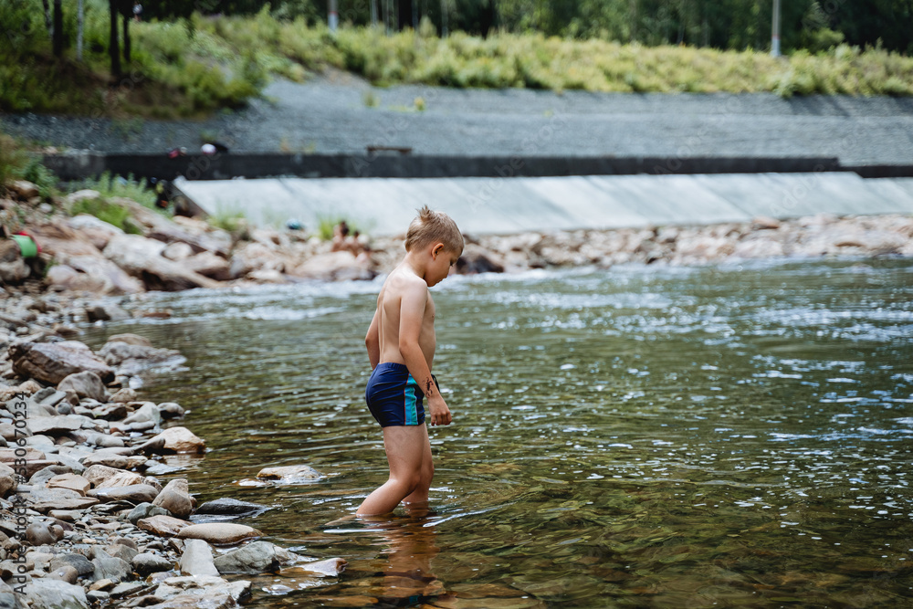 A child enters the water, summer holidays on the river, a boy alone bathes in the cold water of a mountain river, a stone bed, a summer vacation in nature, swimming trunks on the body A child enters the water, summer holidays on the river, a boy alone bathes in the cold water of a mountain river, a stone bed, a summer vacation in nature, swimming trunks on the body