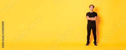 Full length of smiling confident asian blong guy, cross arms chest over yellow background