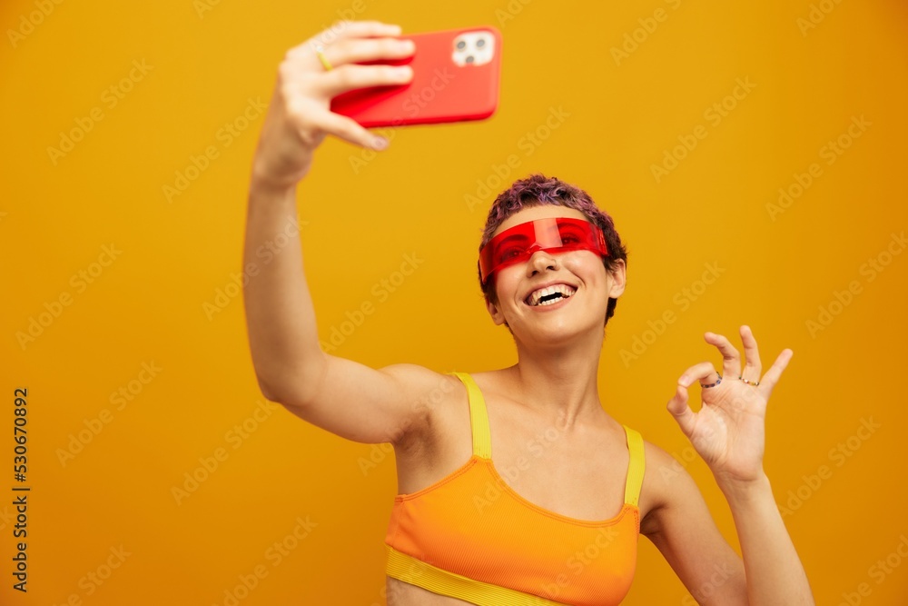 Woman smiles and shows her tongue in unusual glasses millennial takes selfies on her phone in sportswear against an orange studio backdrop, free space Woman smiles and shows her tongue in unusual glasses millennial takes selfies on her phone in sportswear against an orange studio backdrop, free space