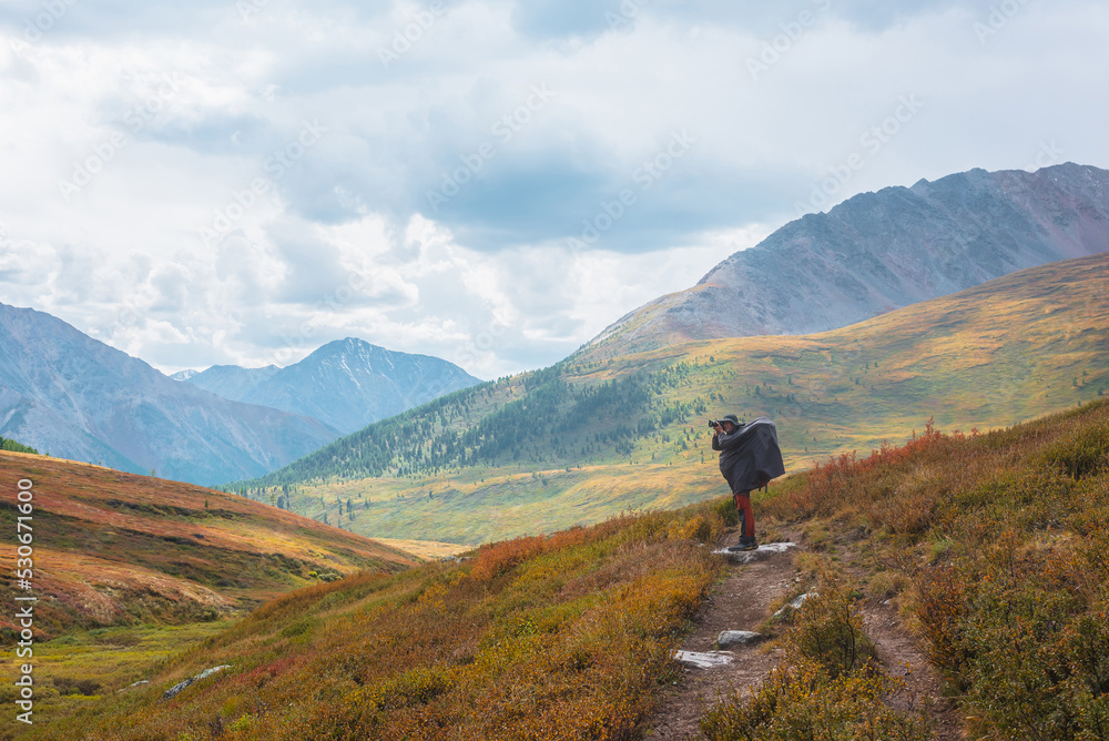 Alone traveler in raincoat with large backpack photographs nature among fading autumn vegetations on hiking trail through mountain pass. Backpacker with photo camera shoots autumn mountain landscape.