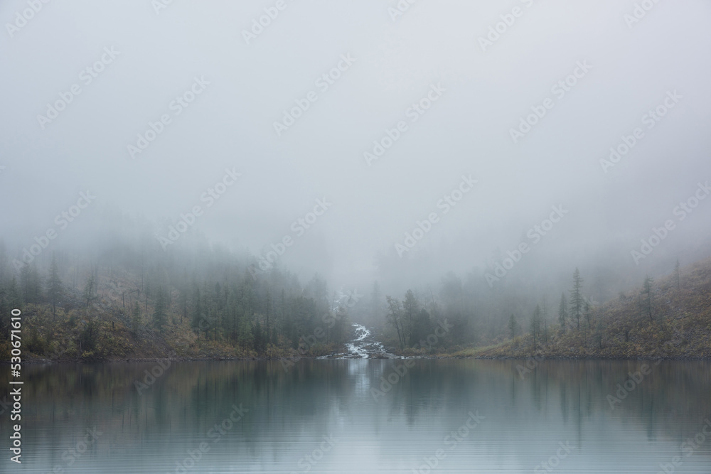 Mountain creek flows from forest hills into glacial lake in mysterious fog. Small river and coniferous trees reflected in calm alpine lake in misty morning. Tranquil landscape in fading autumn colors.