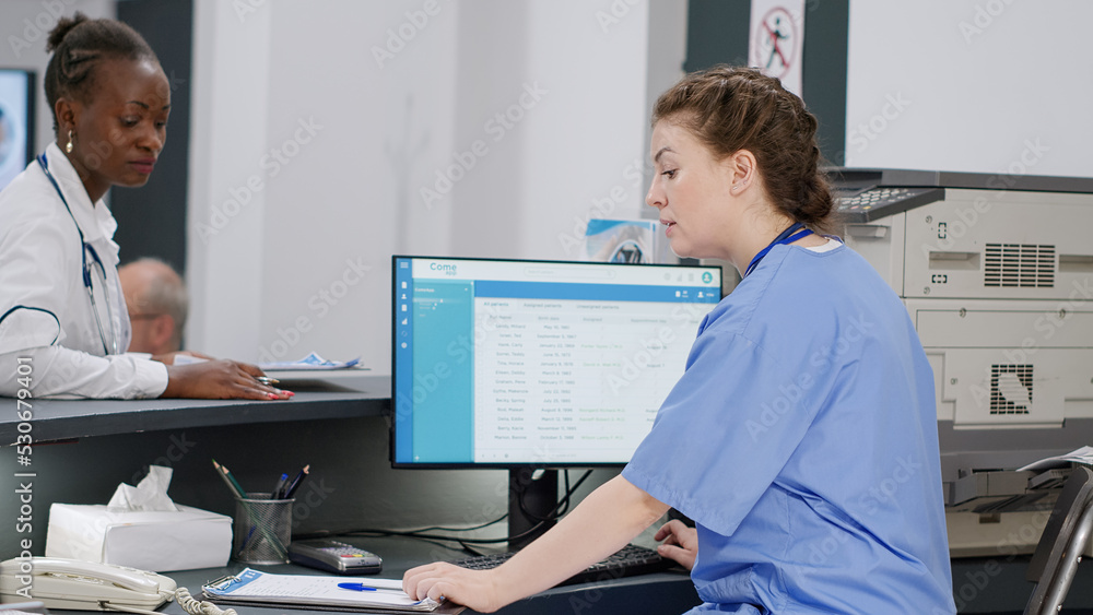 Obraz premium Medical assistant working on patient appointments at reception desk, helping african american doctor with information to do consultation with senior man. Diverse staff at healthcare clinic.