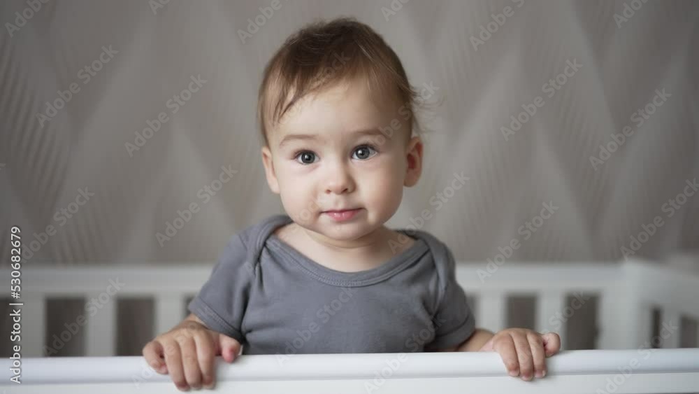 Adorable smiling baby boy standing in white cot swaying a little. Child smiles sweetly showing two lower new teeth. Close up. Adorable smiling baby boy standing in white cot swaying a little. Child smiles sweetly showing two lower new teeth. Close up.