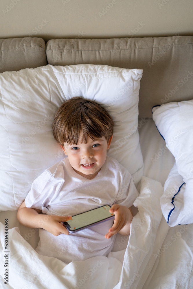 Happy little boy talking on smartphone, looking aside while sitting on bed Happy little boy talking on smartphone, looking aside while sitting on bed